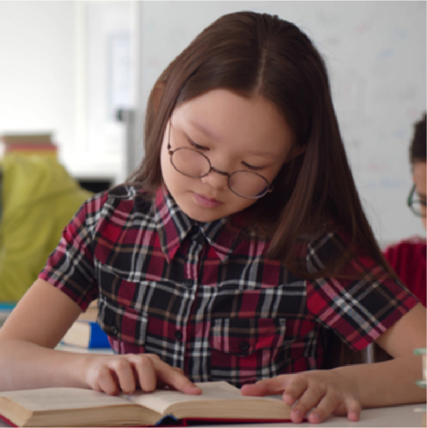 A wise school girl in glasses with a red plaid school uniform reading a book in her hands. Other classmates studying in the background