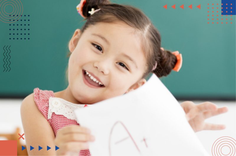 A little girl smiling and holding her grade sheets. With the A+ grade sign on the paper sheet.