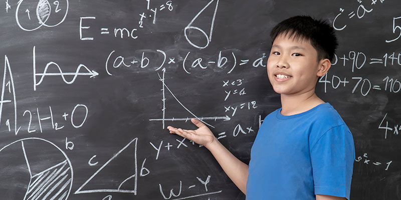 A young boy in a classroom near the blackboard teaches and explains complex math problems.