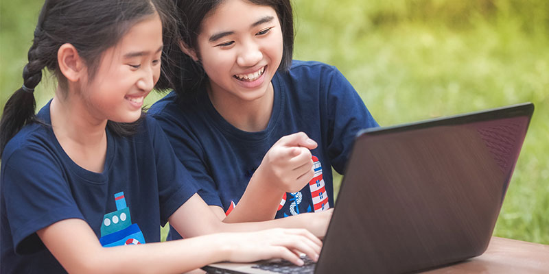 Two young girls are smiling and using a laptop while sitting in a lush open environment.