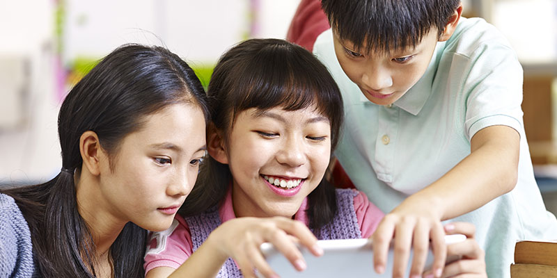 Three happy school students sitting in their classroom smiling and looking at a tablet.