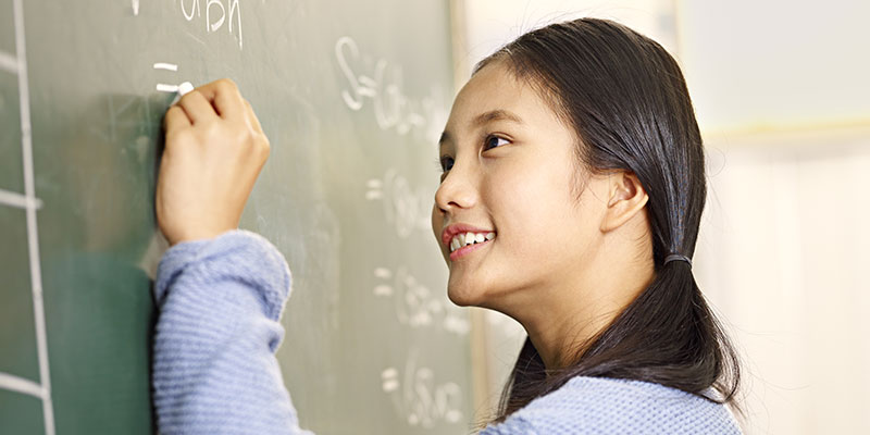 An elementary school girl student writing the answer to the geometrical problem with white chalk on a blackboard