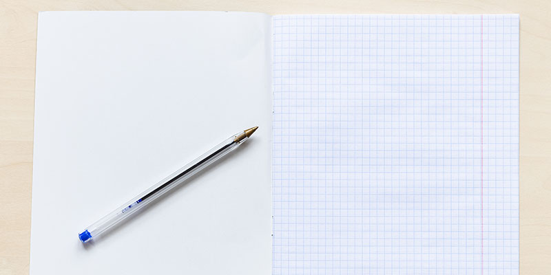 A top view of an open notebook and a pen sits atop a light brown wooden surface.