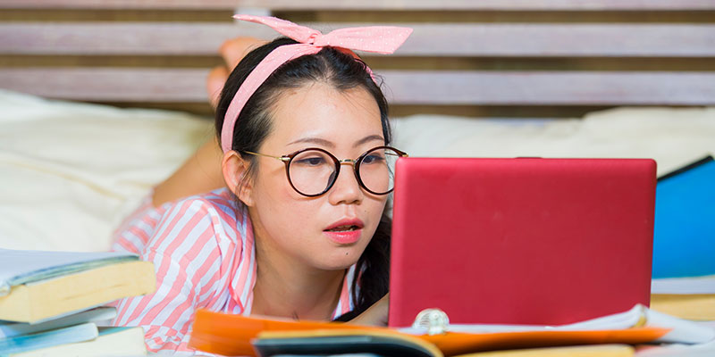 A young girl wearing specs and a pink headband studying in her bedroom with her laptop and books.