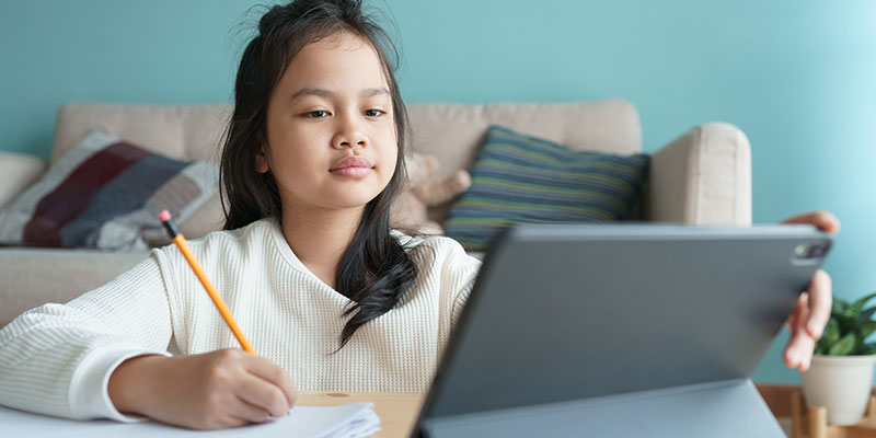 In her home living room, a primary school girl in a white top prepares for her PSLE exam with her tablet computer, notebook, and pencil