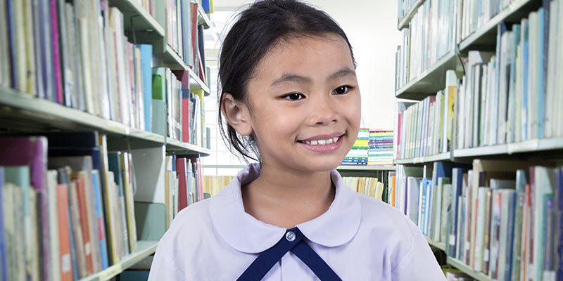 A cute primary school girl smiles while selecting PSLE English composition books in the library.