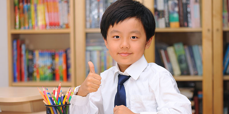 A young primary school boy wearing a white shirt and a tie sitting in a classroom and showing a thumbs up with his hands.