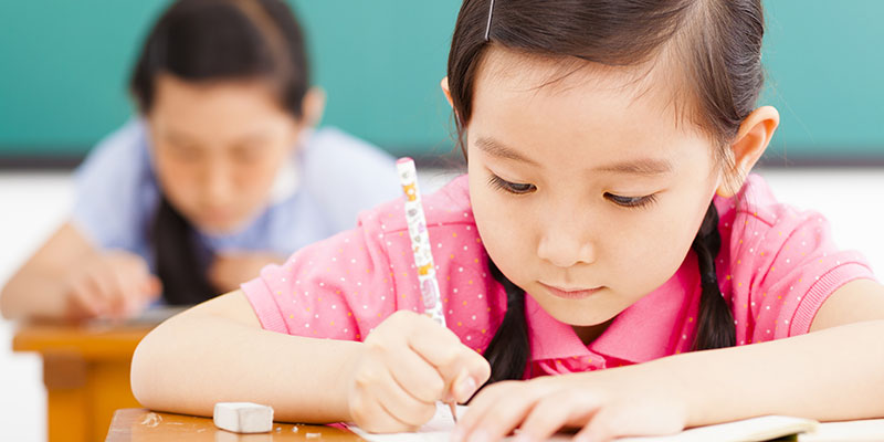 In her classroom, a girl child writes PSLE Situational Writing with other students.