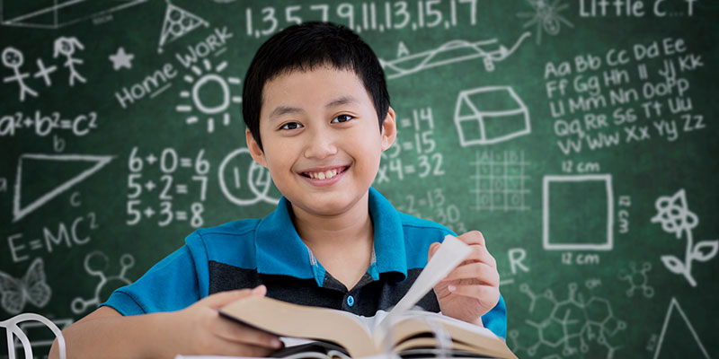 A primary school boy is holding a book in his hand and smiling, with a chalkboard background with creative writings on it.