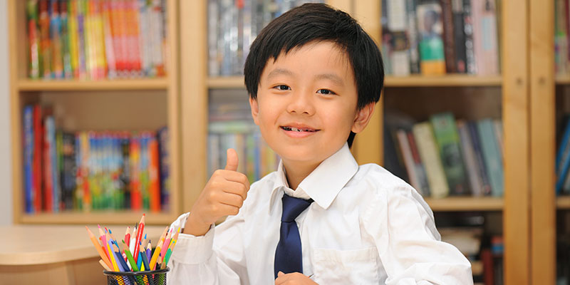 A small schoolboy is gesturing thumbs up with a background of books organized on a shelf.