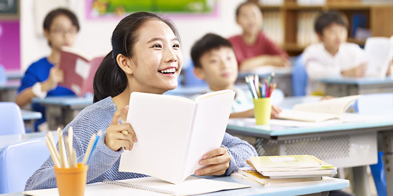 A girl student is holding a notebook in her hand and smiling at her teacher, there are other students behind her listening to the class.