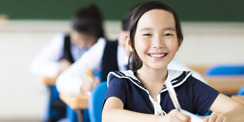 A happy Asian girl seated inside a classroom is writing in a notebook.