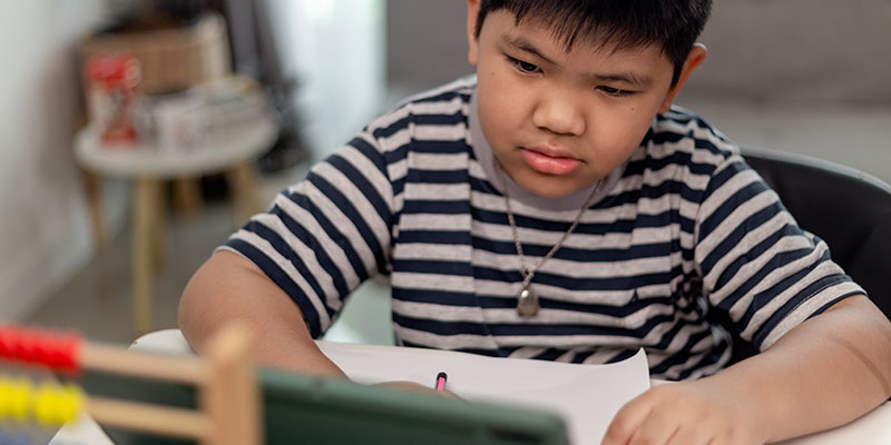 A small boy writes notes in a notebook from a monitor in front of him.