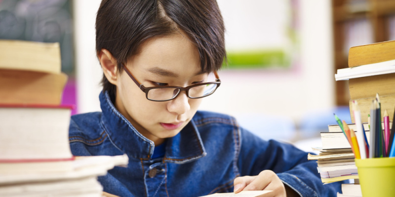 An Asian elementary school boy wearing glasses reading a thick english book.