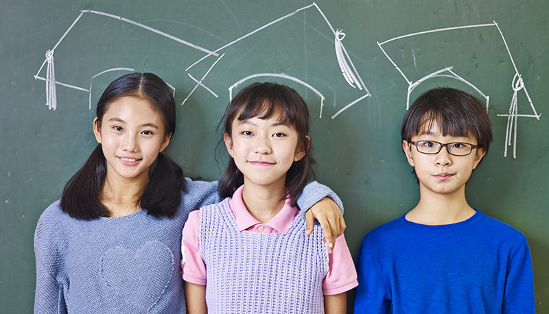 Three Asian primary school children standing in front of a chalkboard underneath chalk-drawn graduation caps illustrate ace PSLE with StudySmart.